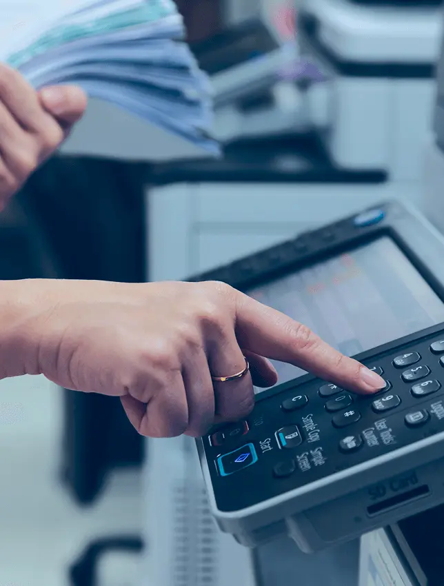 A hand pressing a button on a photocopier. This person is also holding a large stack of papers in their other hand.