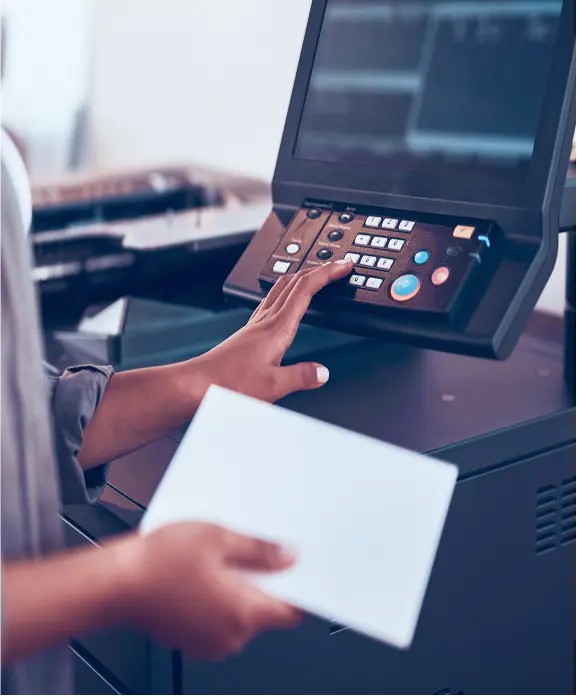 A woman presses buttons on a photocopier while holding a sheet of paper.